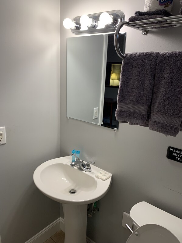 A bathroom with a white sink, a mirror with lights, a towel rack, and a sign on the wall.