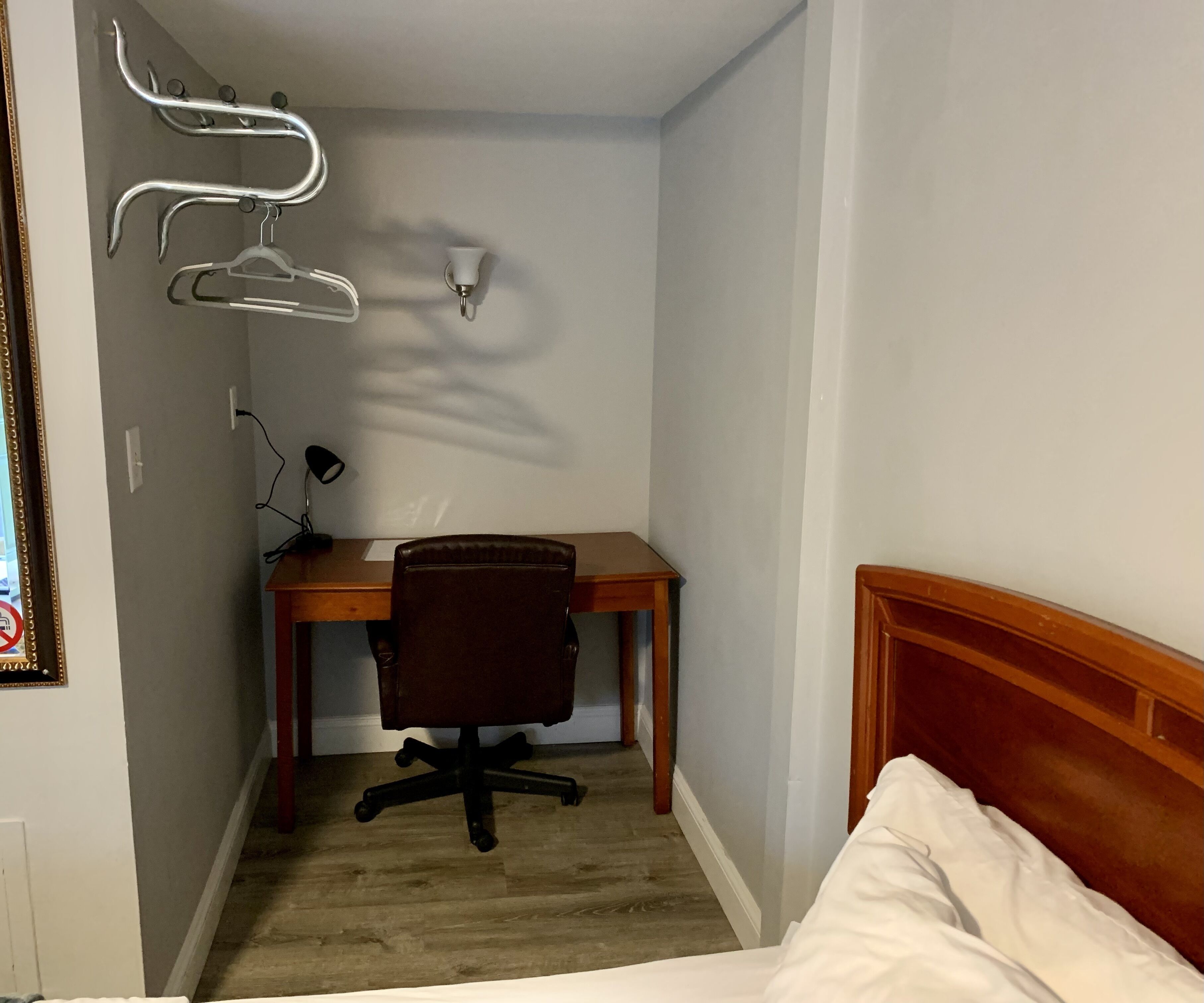 A small, neatly arranged room with a wooden desk, a black office chair, a bed with white bedding, and a wall-mounted lamp.