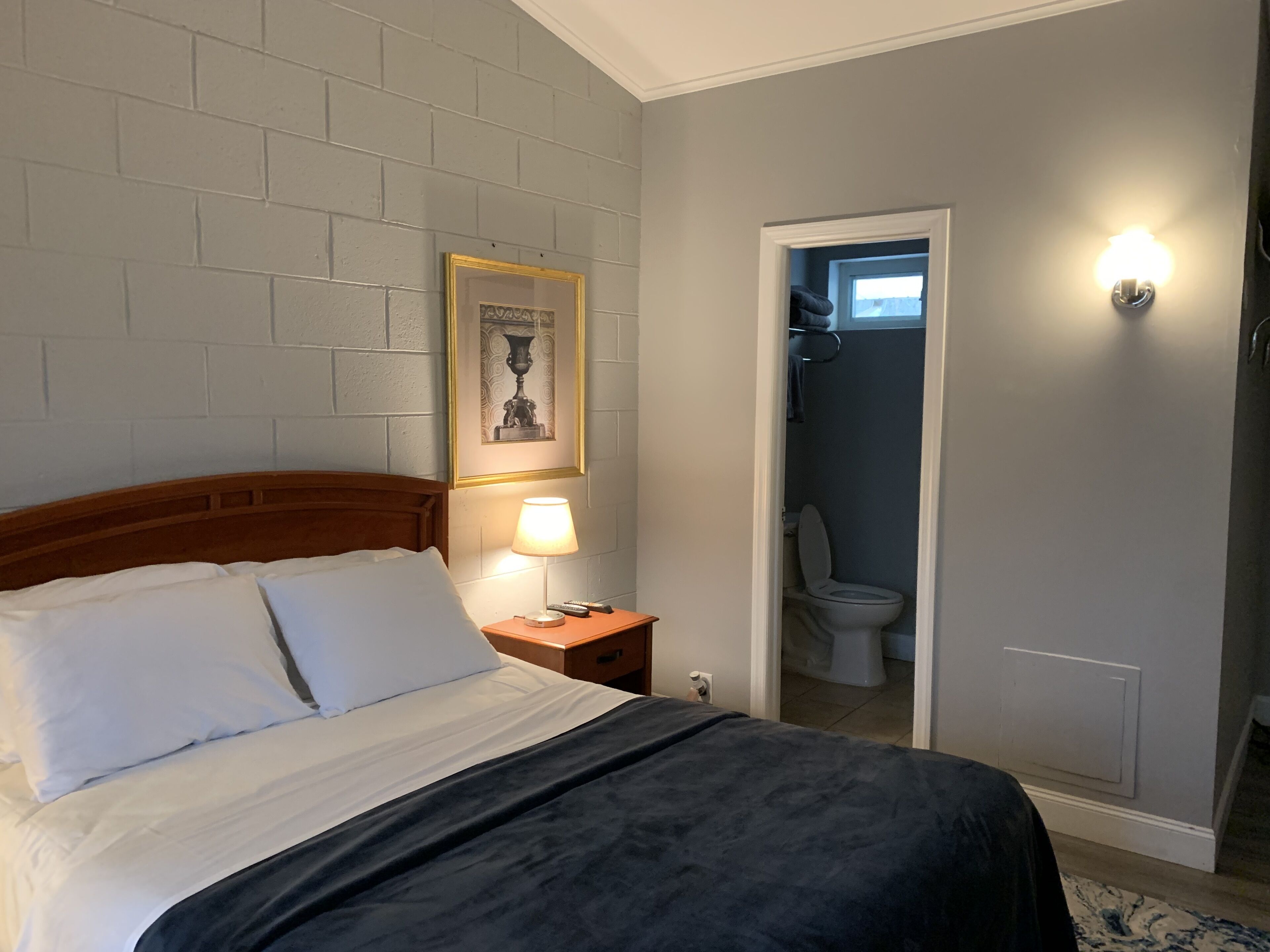 Ein Hotelzimmer mit einem Bett, einem Nachttisch mit Lampe, einem gerahmten Bild an der Wand und einer Tür zu einem Badezimmer.