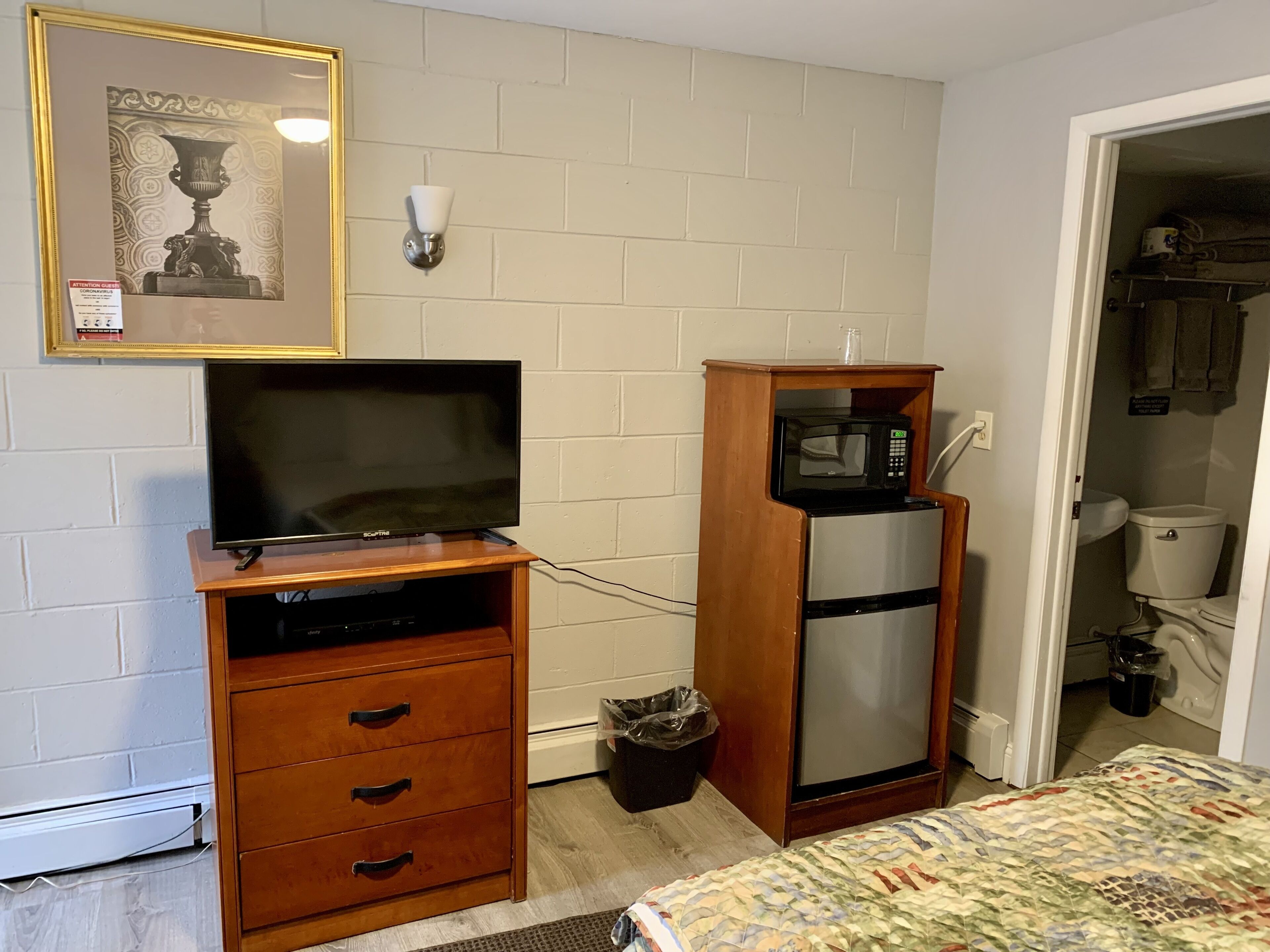 Una habitación pequeña con televisión sobre un soporte de madera, microondas en un estante de madera y cama con manta estampada.