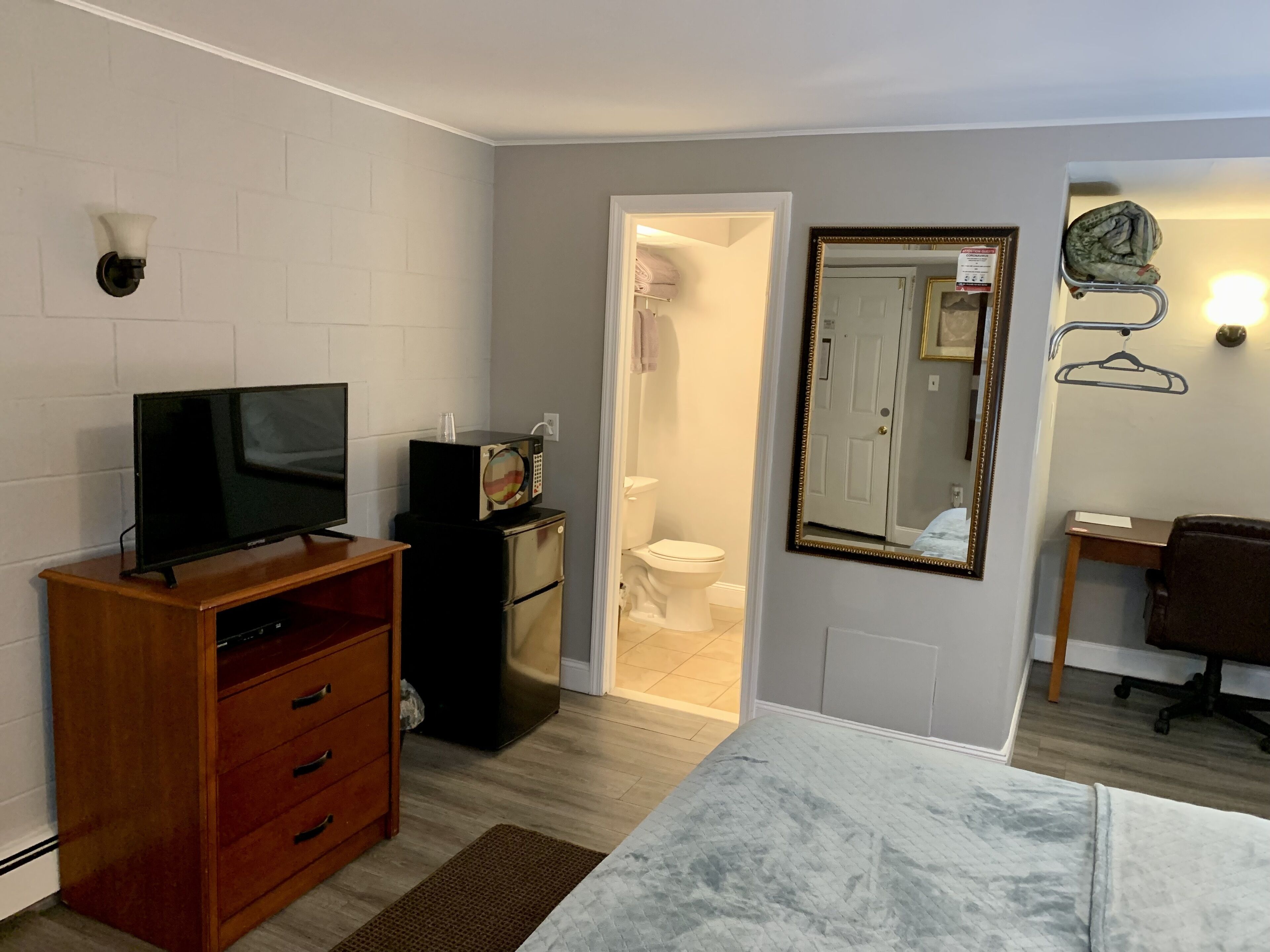 A compact room with a television on a wooden stand, a small black refrigerator, a mirror with a gold frame, and a chair.