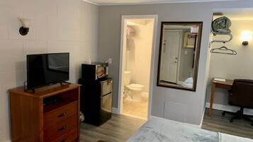 A compact room with a television on a wooden stand, a small black refrigerator, a mirror with a gold frame, and a chair.