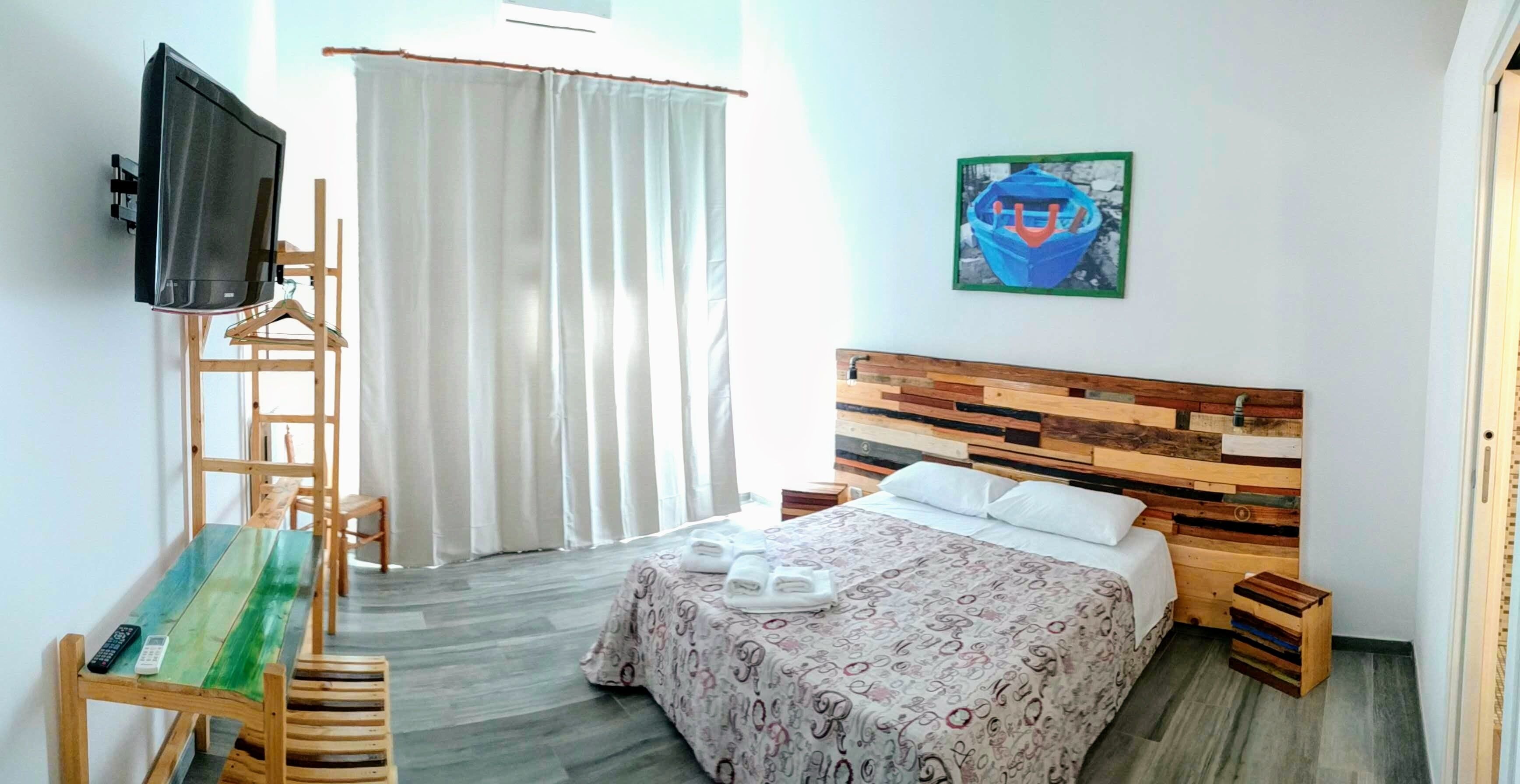 A hotel room with a bed, a TV mounted on the wall, a wooden headboard, a bench, and a painting on the wall.