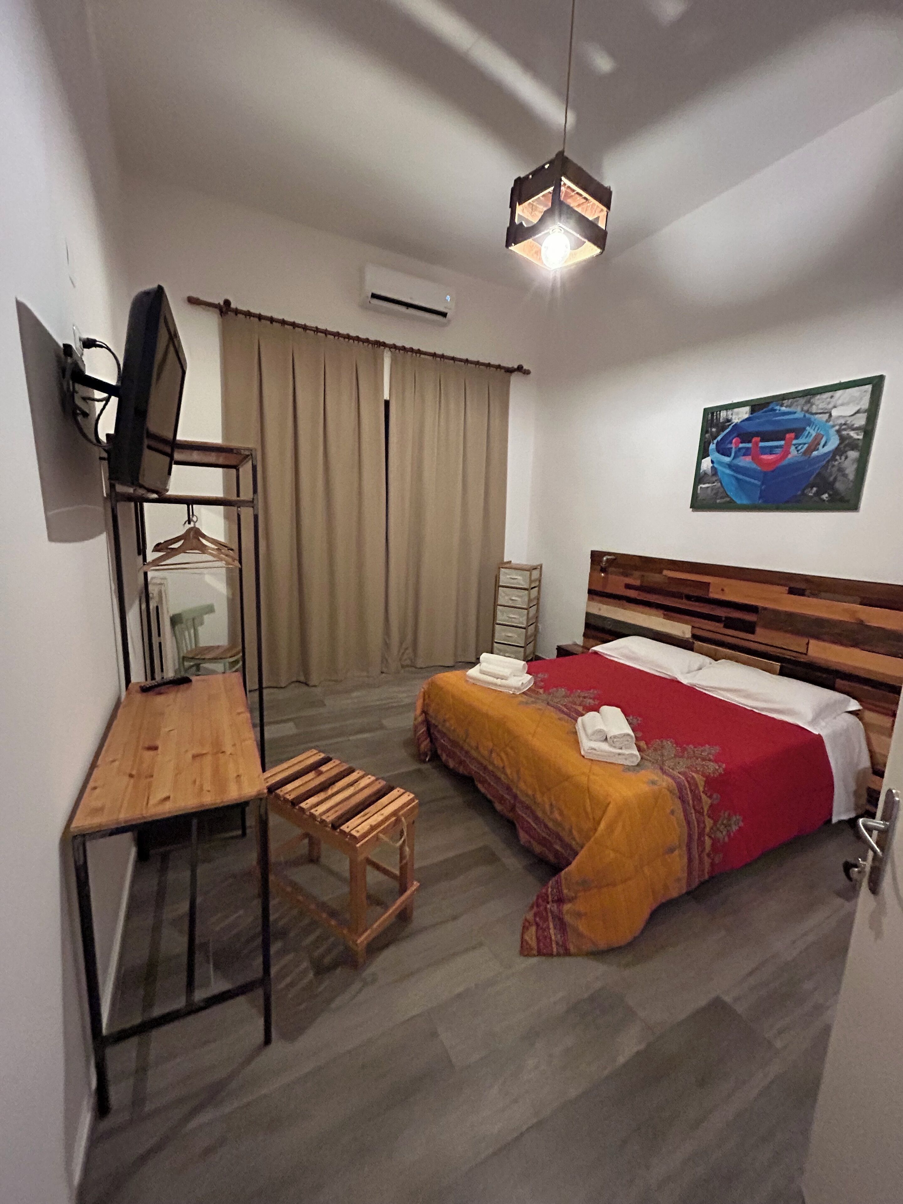 Une chambre d’hôtel de taille réduite, comprenant un lit simple, un bureau, une chaise, un petit tabouret, un téléviseur et un tableau au mur.