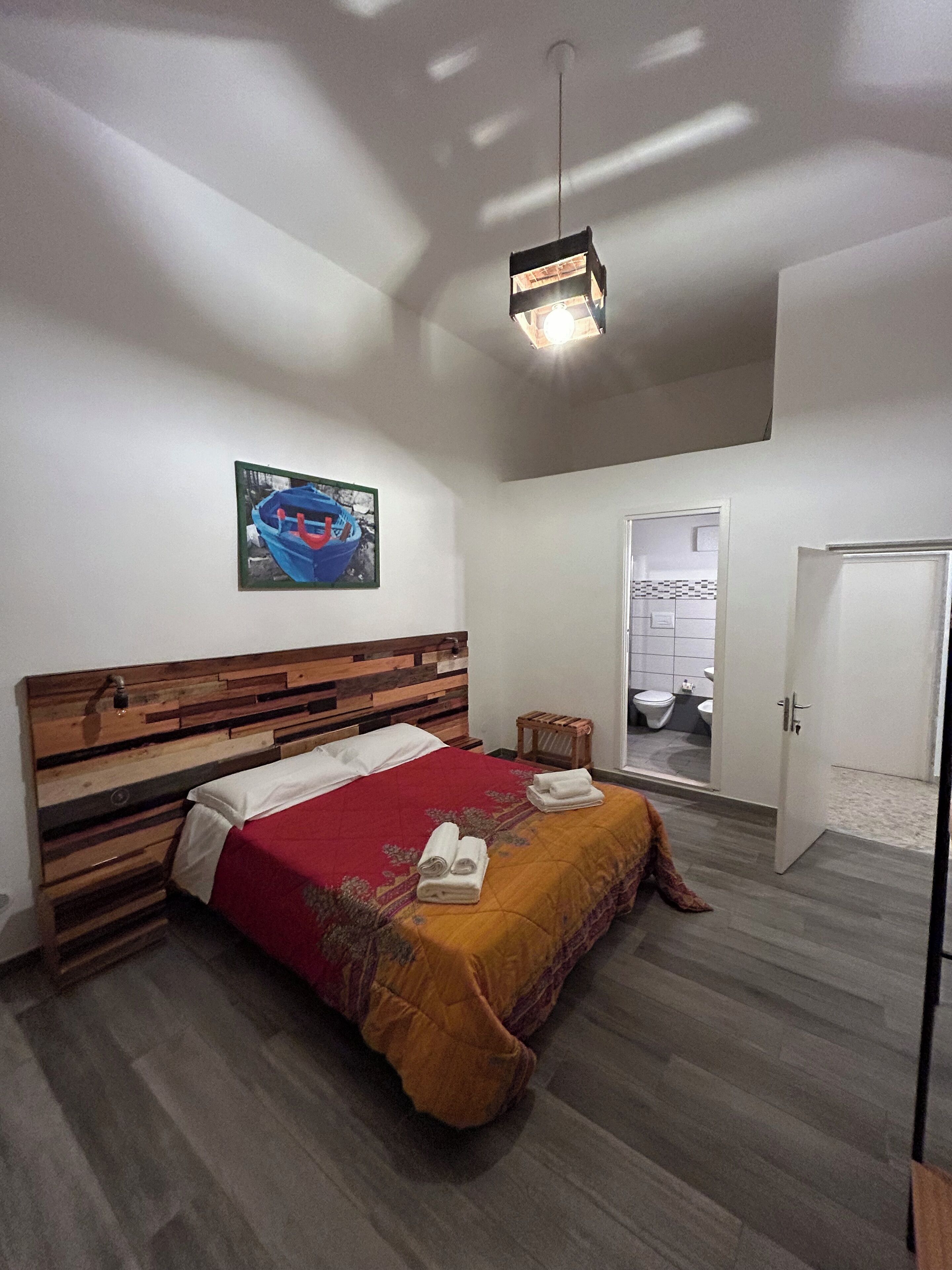 Un dormitorio con cabecera de madera, cama con colcha roja y amarilla, un taburete de madera pequeño y una puerta que da al baño.