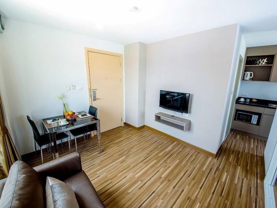 A modern living room with a wooden floor, a brown leather sofa, a dining table with chairs, a TV mounted on the wall, and a kitchenette with a microwave and toaster.