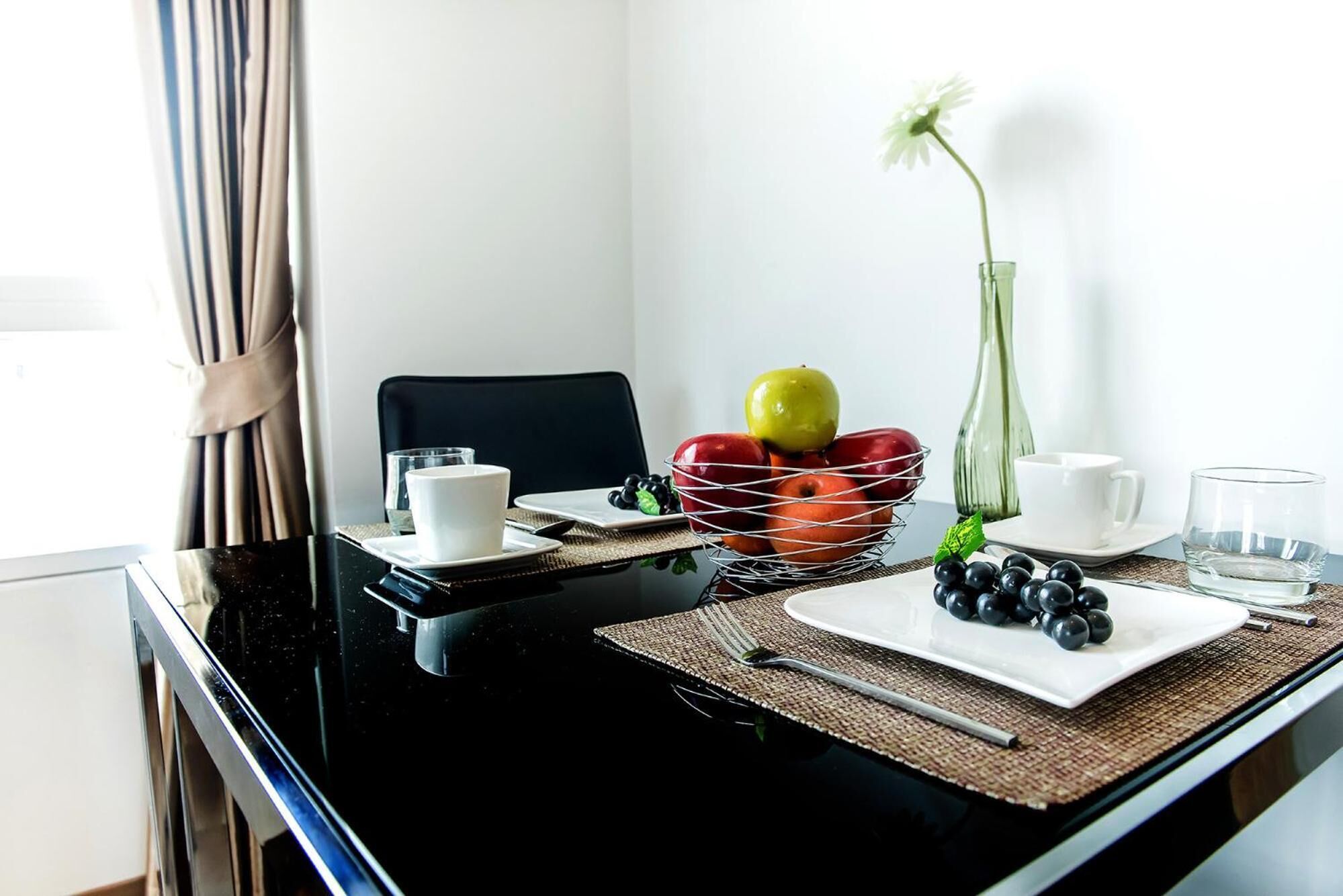 A dining area with a black table, white plates, a fruit basket, and a vase with a single flower.