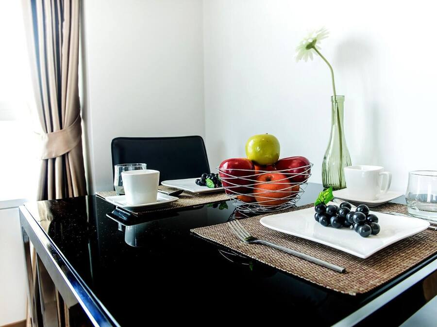 A dining area with a black table, white plates, a fruit basket, and a vase with a single flower.