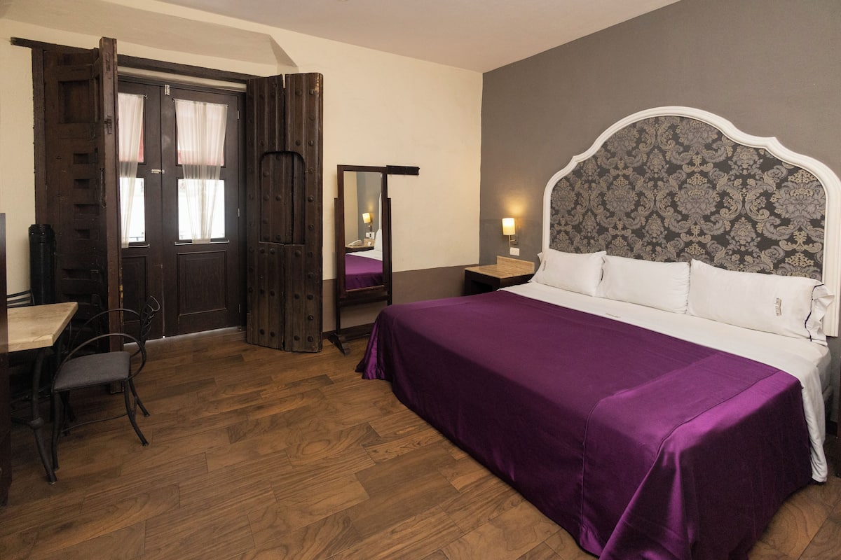Ein Schlafzimmer mit Holzboden, einem Bett mit violetter Bettdecke, einem Holztisch mit Stuhl, einem Spiegel und einem Nachttisch mit Lampe.