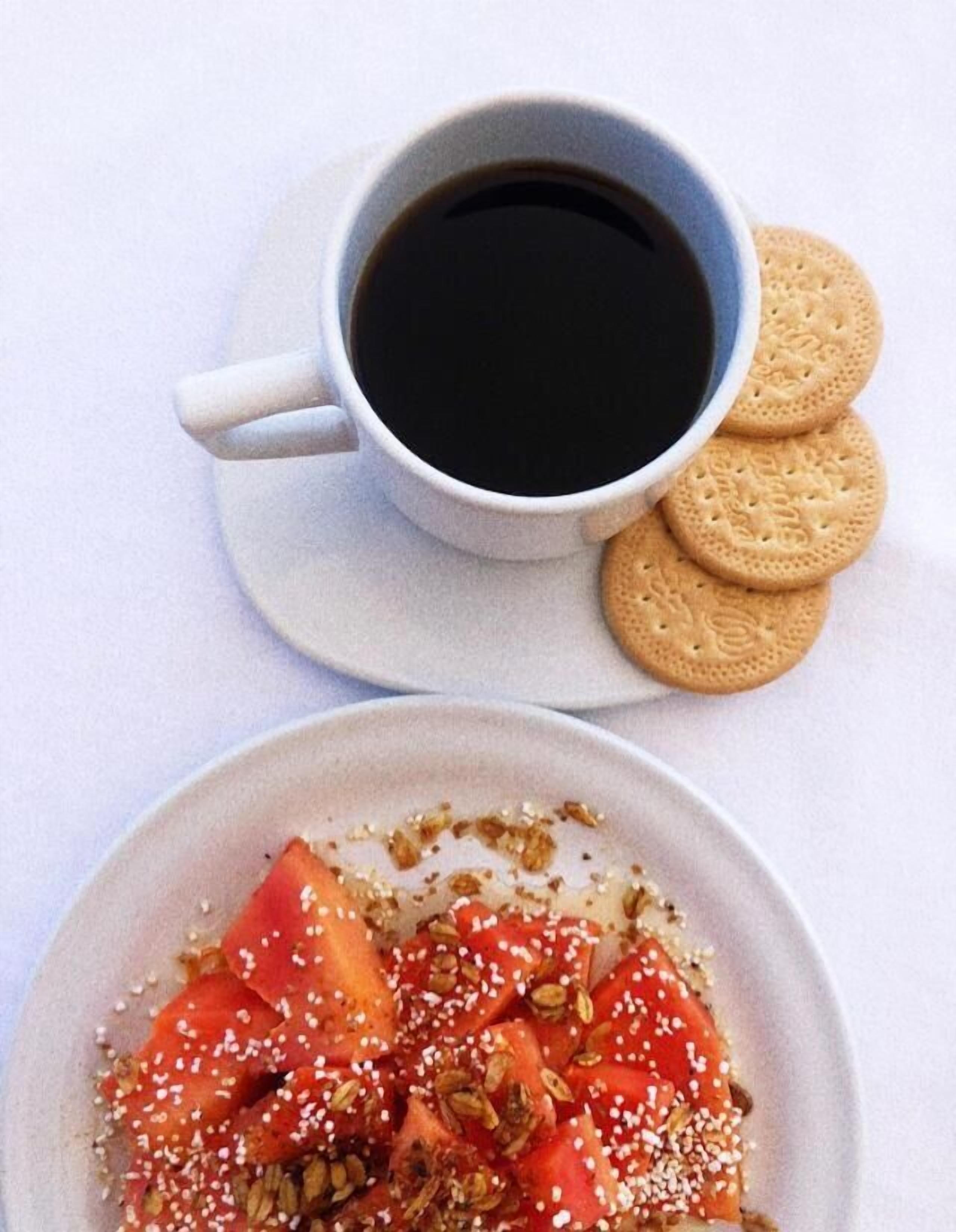 A cup of black coffee, a plate with sliced strawberries and seeds, and a biscuit.
