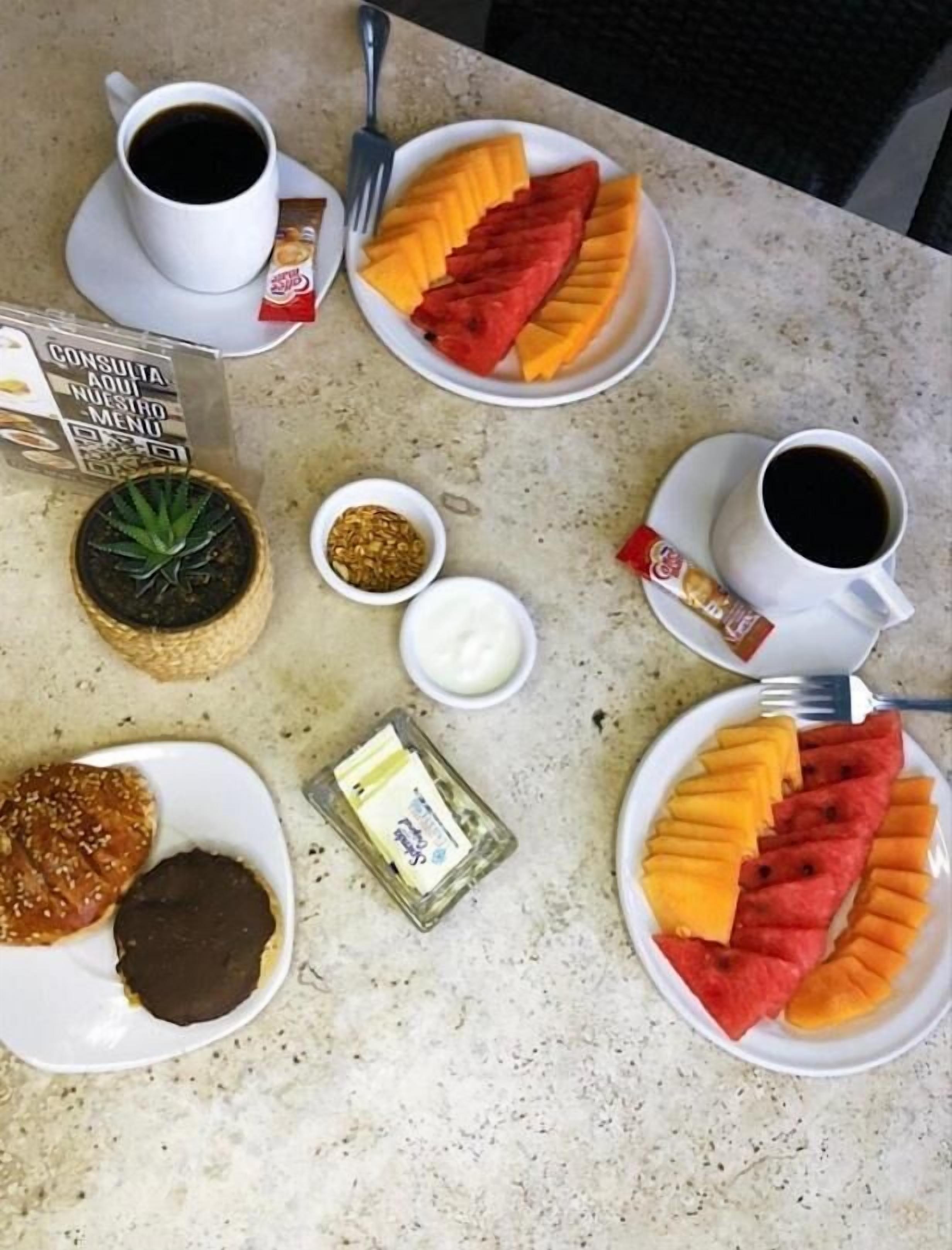 A table with a meal consisting of sliced watermelon, mango, a small plant, a bowl of seeds, a container of butter, a chocolate-covered item, and two cups of coffee.