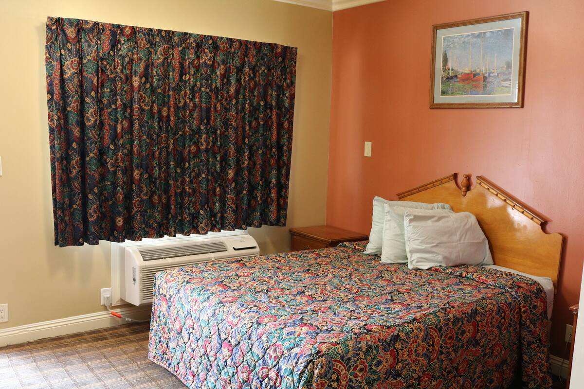 A bedroom with a bed, a window with curtains, a painting on the wall, and an air conditioning unit.