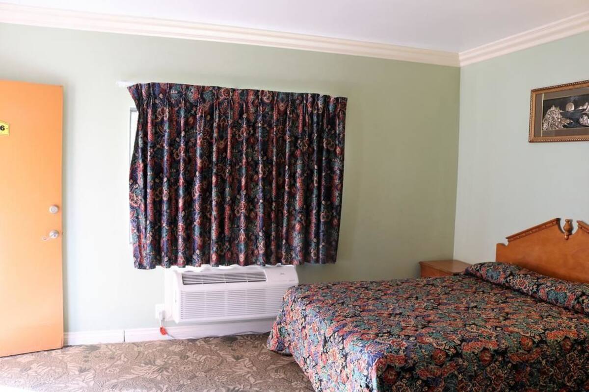 A hotel room with a bed, an air conditioning unit, a door, and a window with patterned curtains.