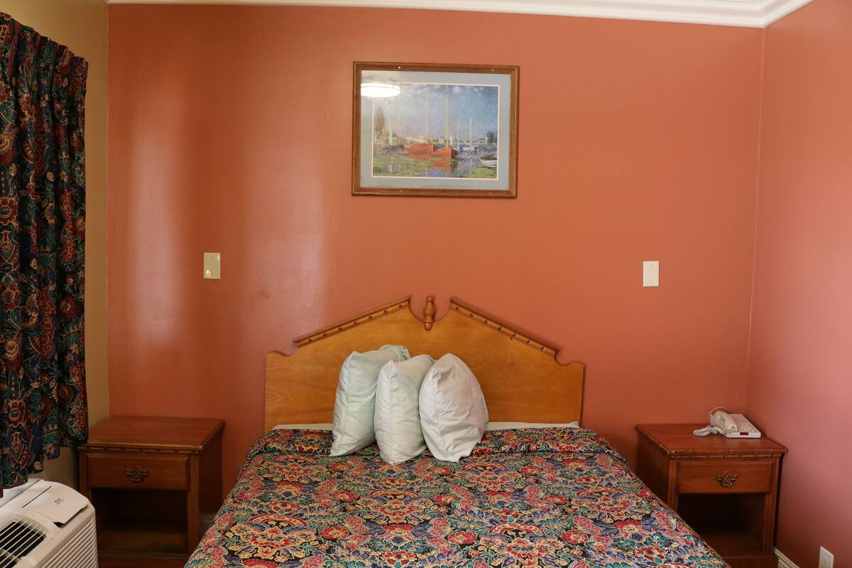 A bedroom with a wooden headboard, a patterned bedspread, two bedside tables, a telephone, and a wall-mounted picture.