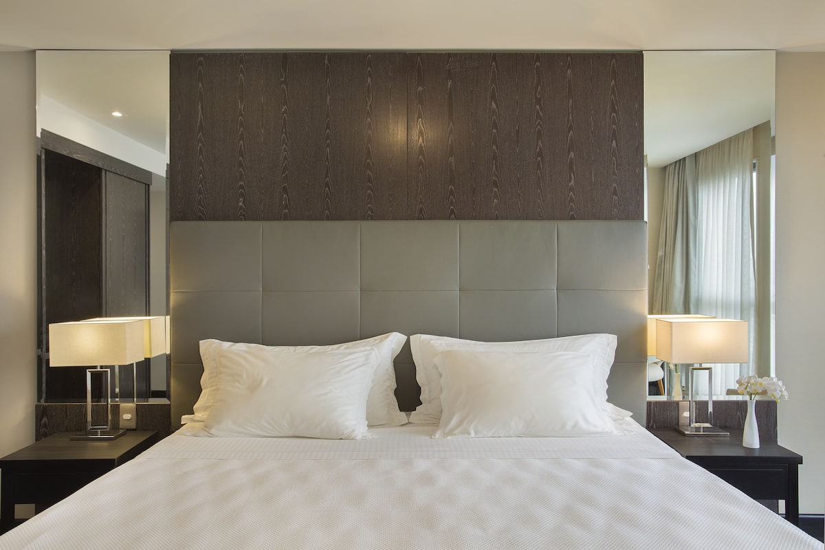 Een moderne hotelkamer met een groot bed, twee nachtkastjes met lampen, een grote spiegel en een hoofdeinde van donker hout.