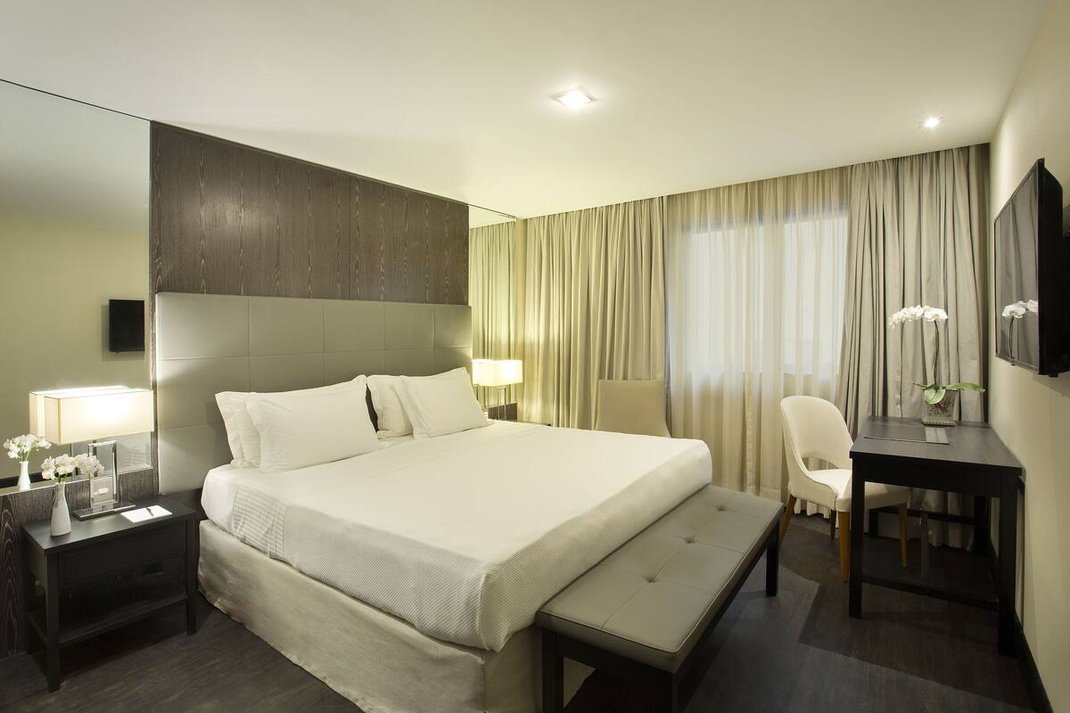A hotel room with a large bed, bedside tables, a desk, a chair, a bench, and a mirror.