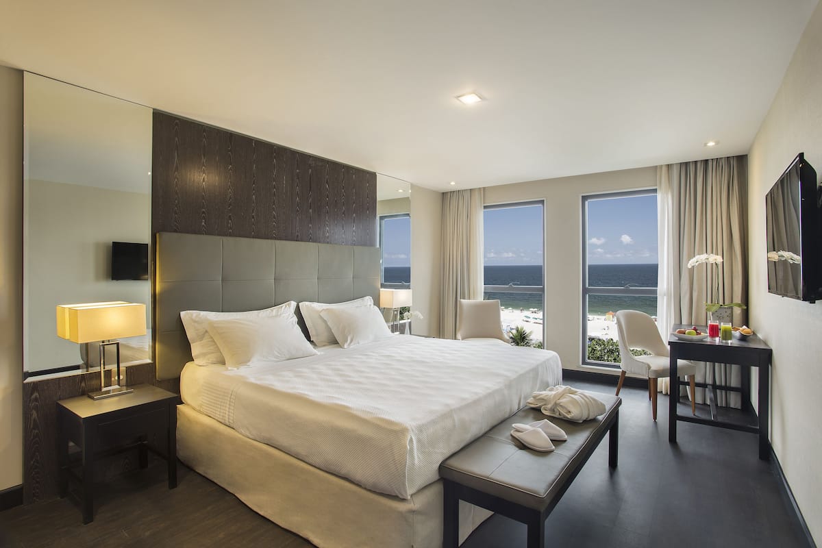 Een hotelkamer met een bed, nachtkastje, lamp, stoel, bureau en een groot raam met uitzicht op de oceaan.