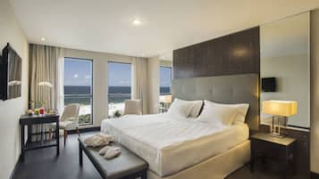 Quarto de hotel com uma cama grande, uma mesa com cadeira, uma mesinha com banqueta e uma televisão sobre um suporte.