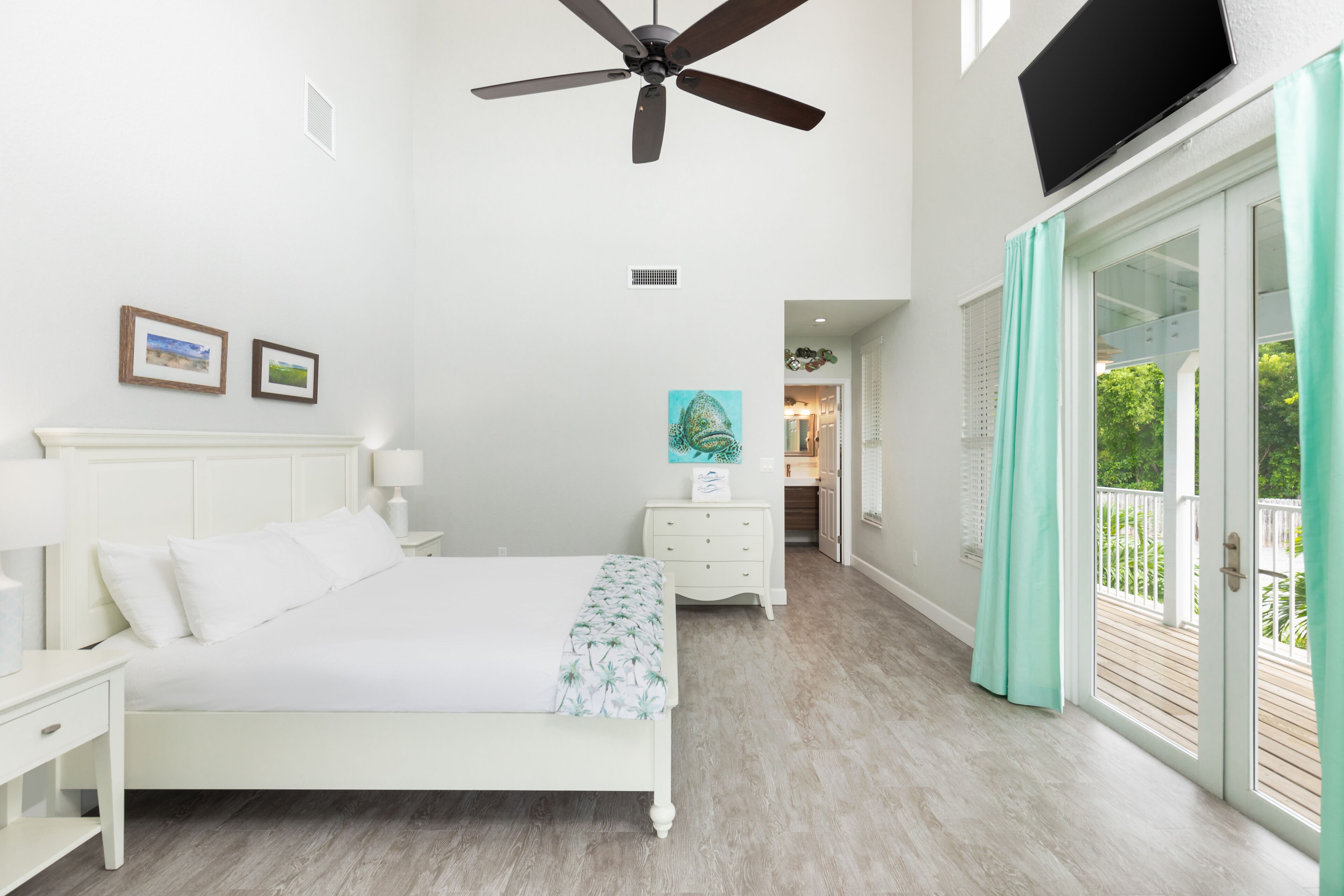A bedroom with a bed, nightstands, a ceiling fan, a painting, and a view of the outdoors through a sliding glass door.