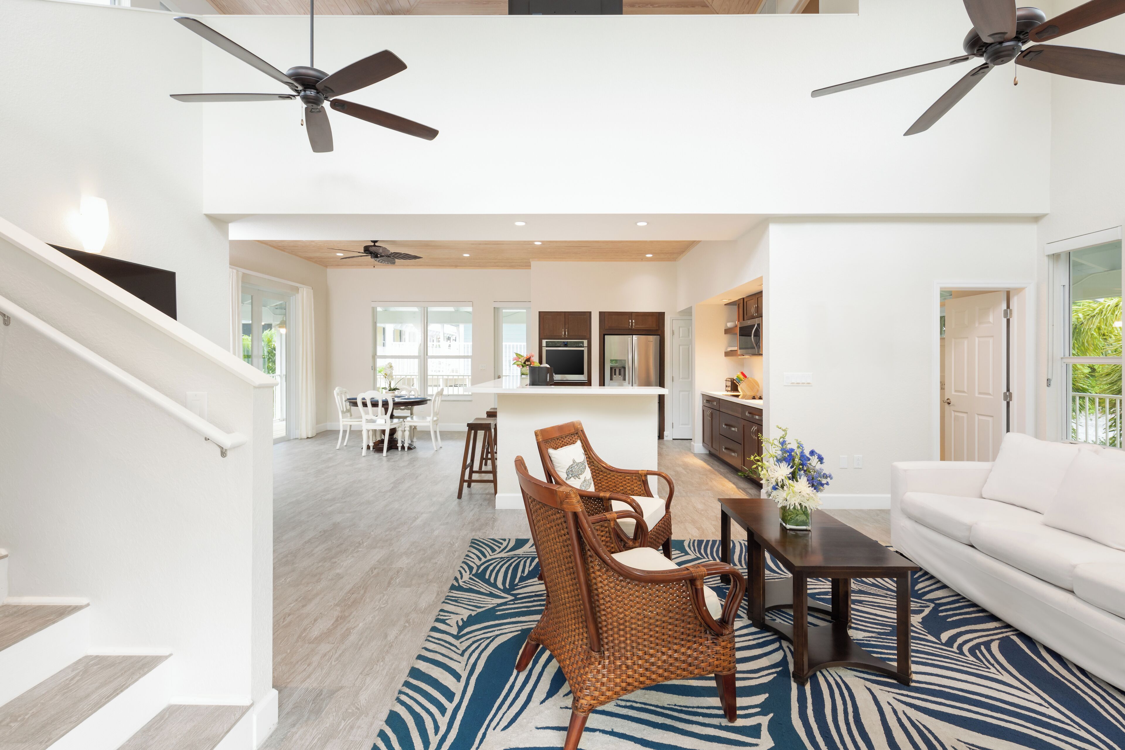 A spacious living area with a high ceiling, two ceiling fans, a dining area with a white table and chairs, a kitchen with modern appliances, and a white sofa.