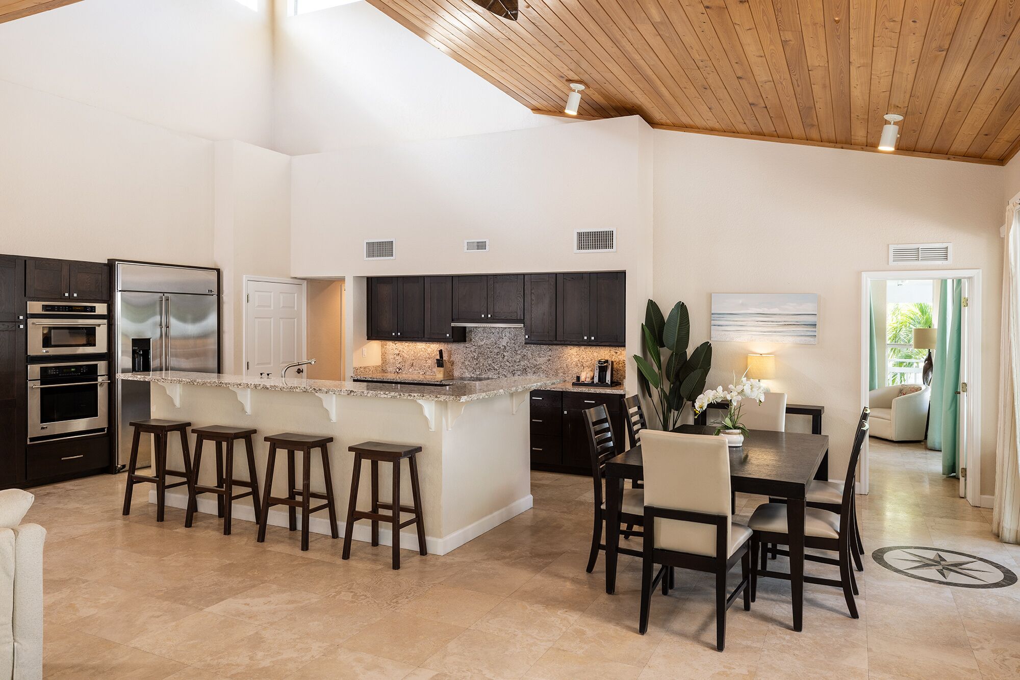 A spacious kitchen with dark cabinets, a granite countertop, and a central island with stools. There are built-in ovens and a refrigerator.
