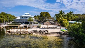 Propiedad frente al mar con un edificio grande, muelle, sillas de descanso y una zona con techo de paja.