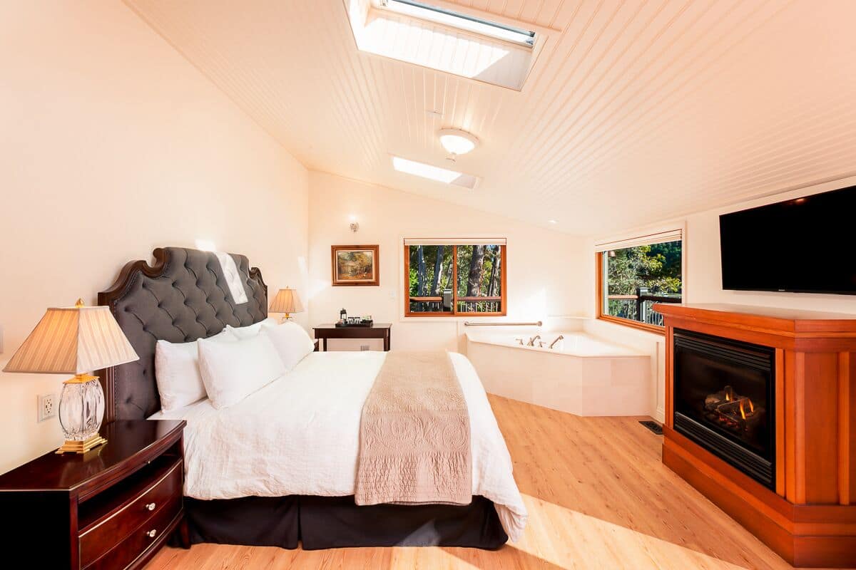A bedroom with a bed, bedside table, lamp, artwork, window, and fireplace.