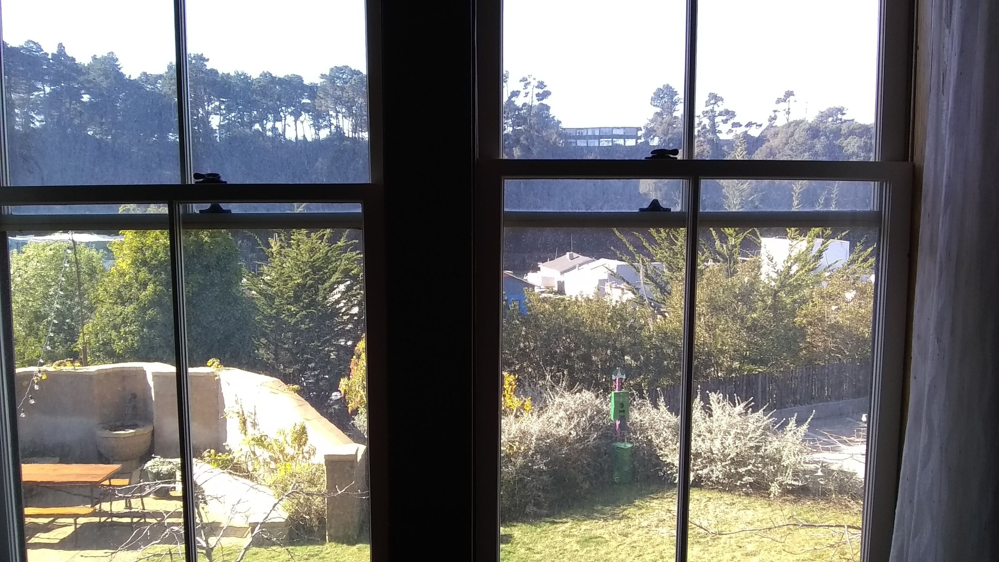 Une vue à travers une fenêtre à carreaux multiples, donnant sur un jardin aménagé avec un barbecue, un mur en pierre et diverses plantes.