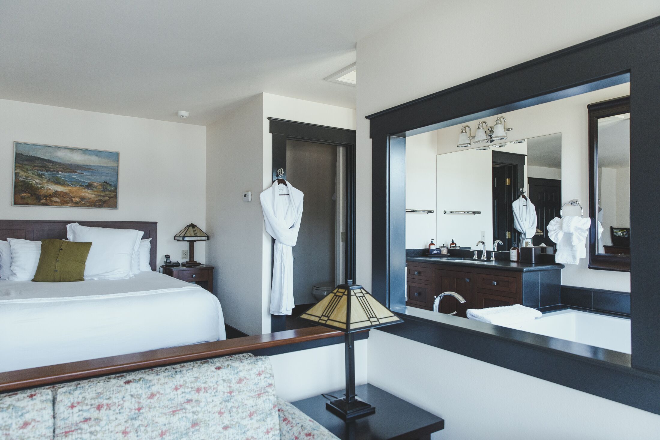 Une chambre d’hôtel comprenant un lit, une table de chevet, une lampe, un miroir et une salle de bain avec un lavabo et un miroir.