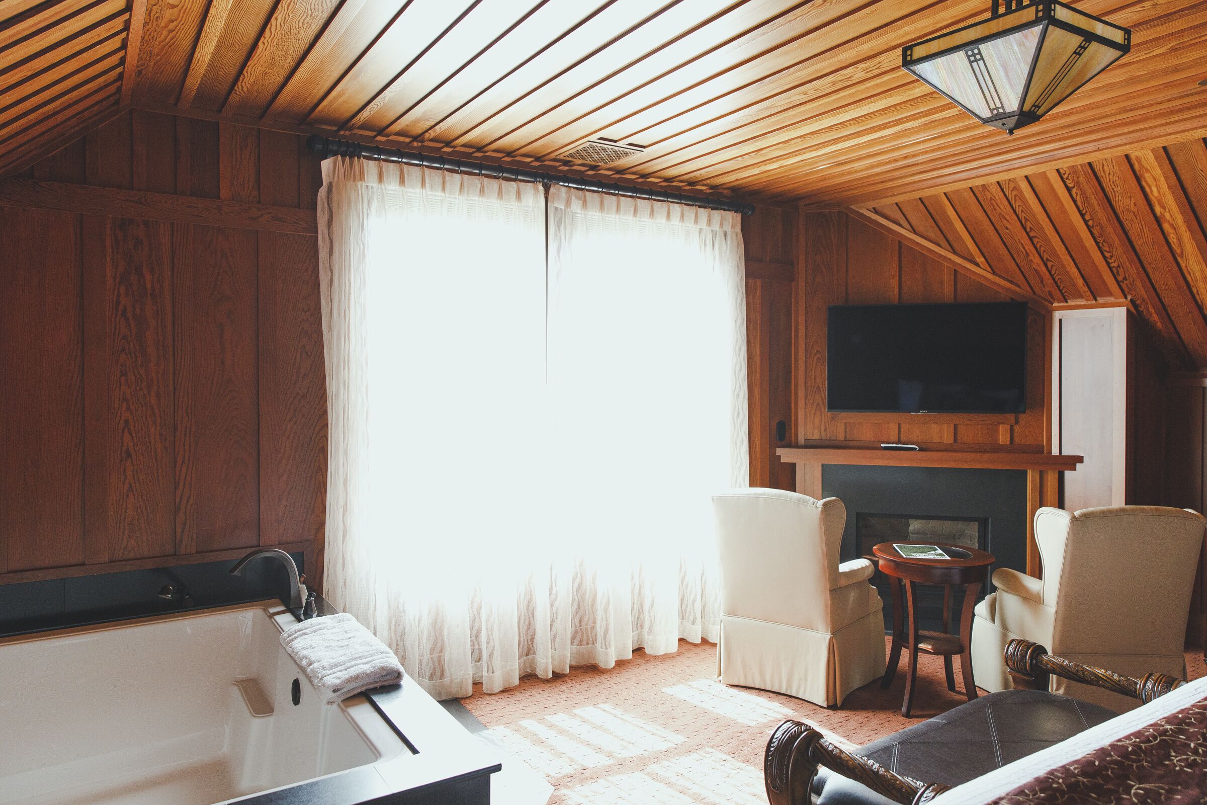 A wooden-paneled room with a bathtub, two chairs, a small table, and a fireplace.