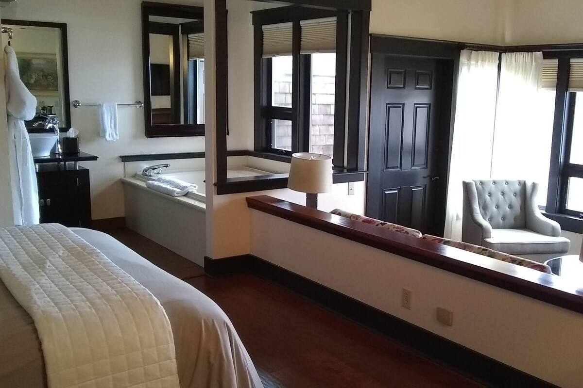 A hotel room with a large bed, a bathroom with a bathtub, a mirror, and a chair by the window.