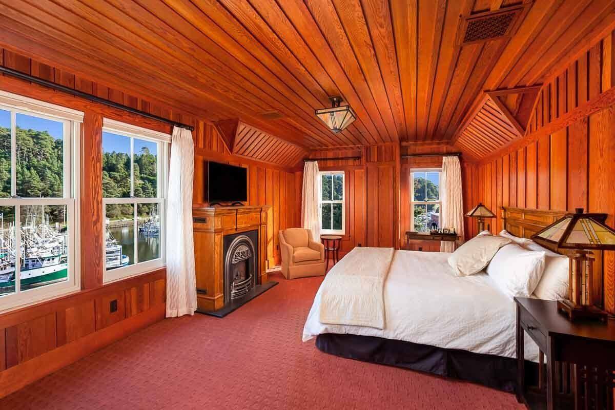 A wooden-paneled bedroom with a bed, a fireplace, a TV, and a view of boats through large windows.