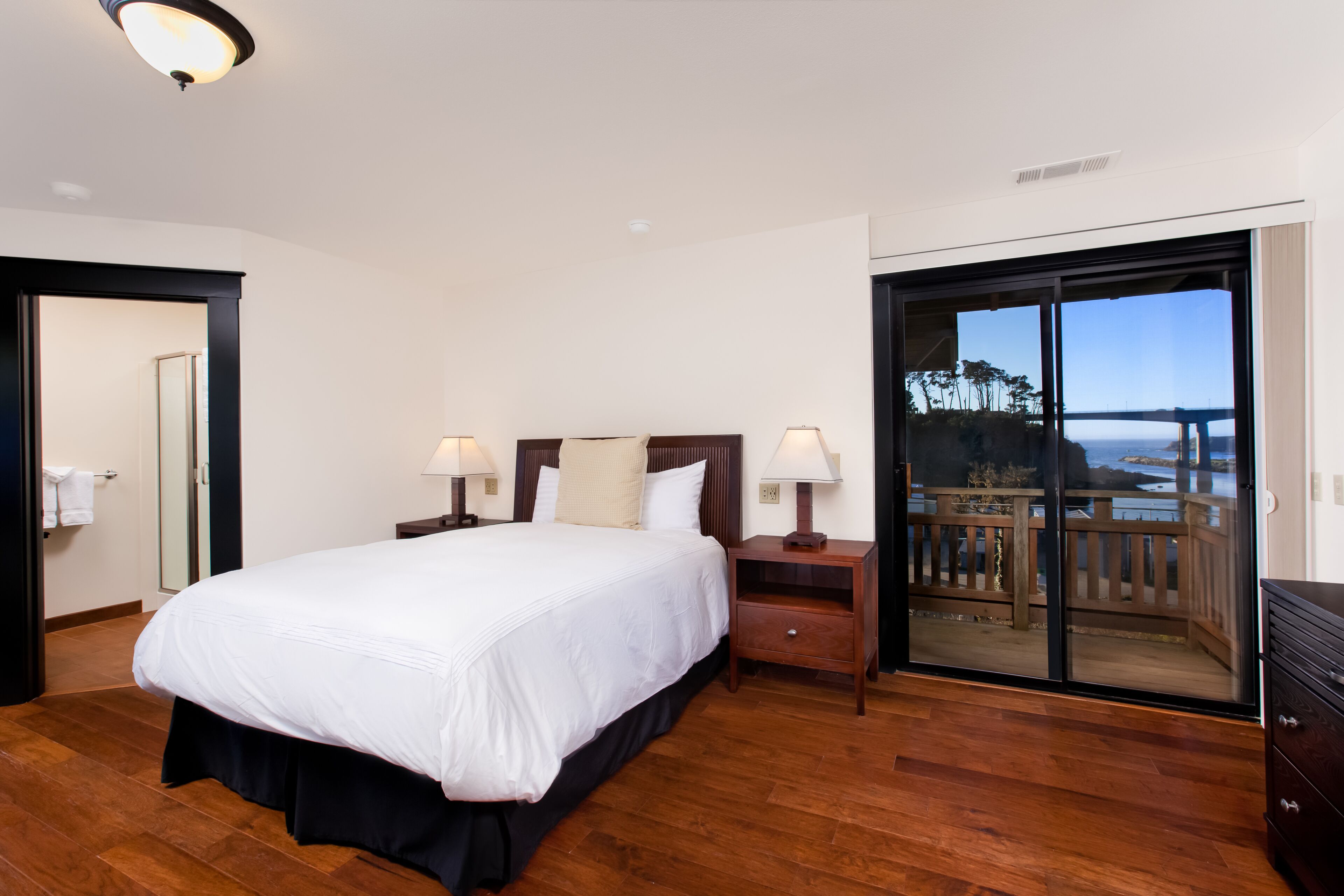 A bedroom with a large bed, two bedside tables with lamps, a wooden floor, and a sliding glass door leading to a balcony with a view.