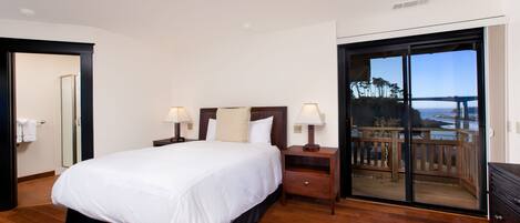 A bedroom with a large bed, two bedside tables with lamps, a wooden floor, and a sliding glass door leading to a balcony with a view.
