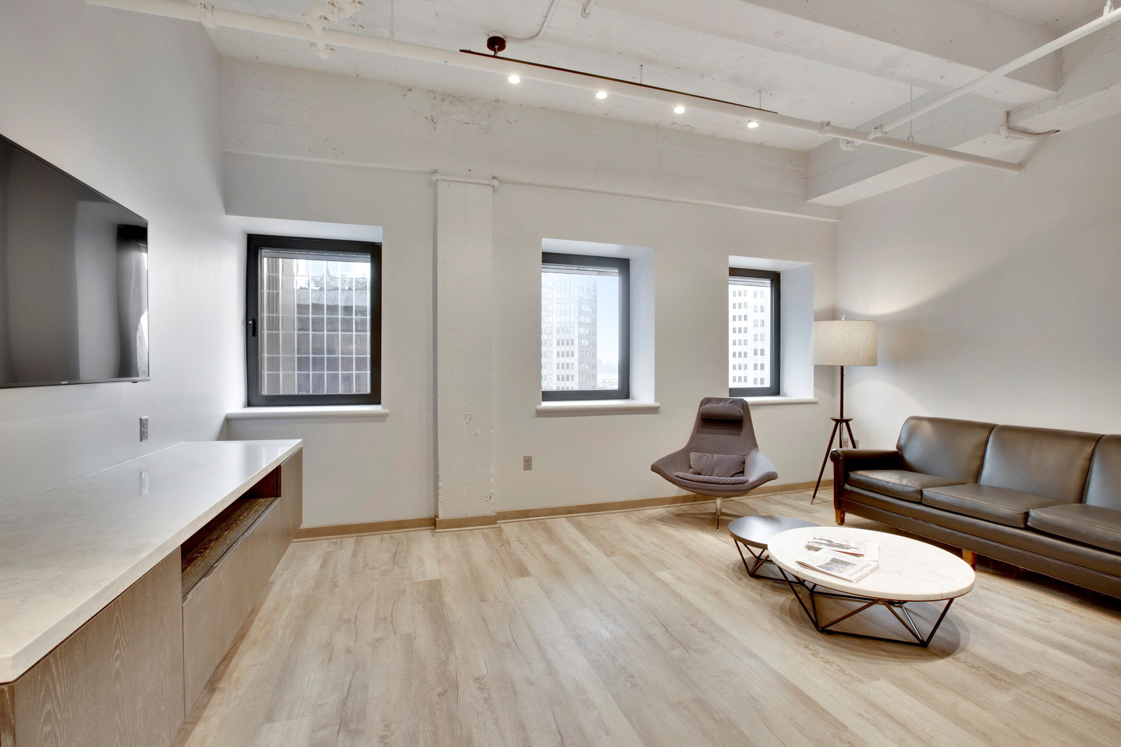 A modern living room with a wooden floor, a round coffee table, a sofa, and a chair. There are three windows providing natural light.