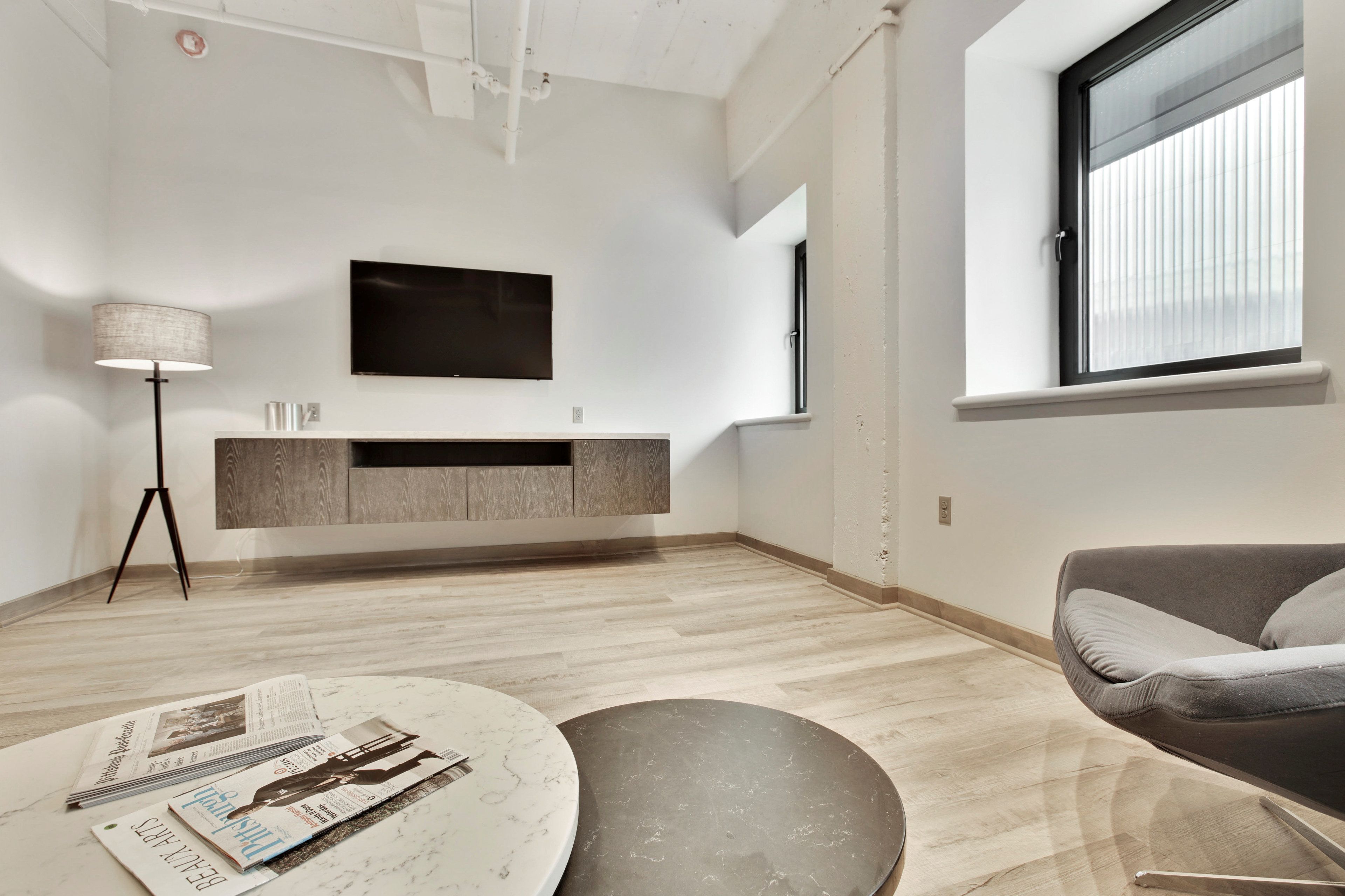 A modern living room with a wooden floor, a round coffee table with magazines, a grey sofa, a TV mounted on the wall, and a window with blinds.