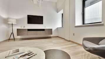 A modern living room with a wooden floor, a round coffee table with magazines, a grey sofa, a TV mounted on the wall, and a window with blinds.