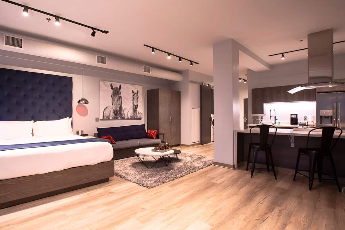 A modern hotel room with a large bed, a sofa, a dining area, and a kitchenette.