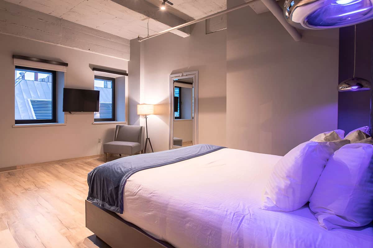 A modern bedroom with a large bed, a chair, a desk, and a TV on the wall.