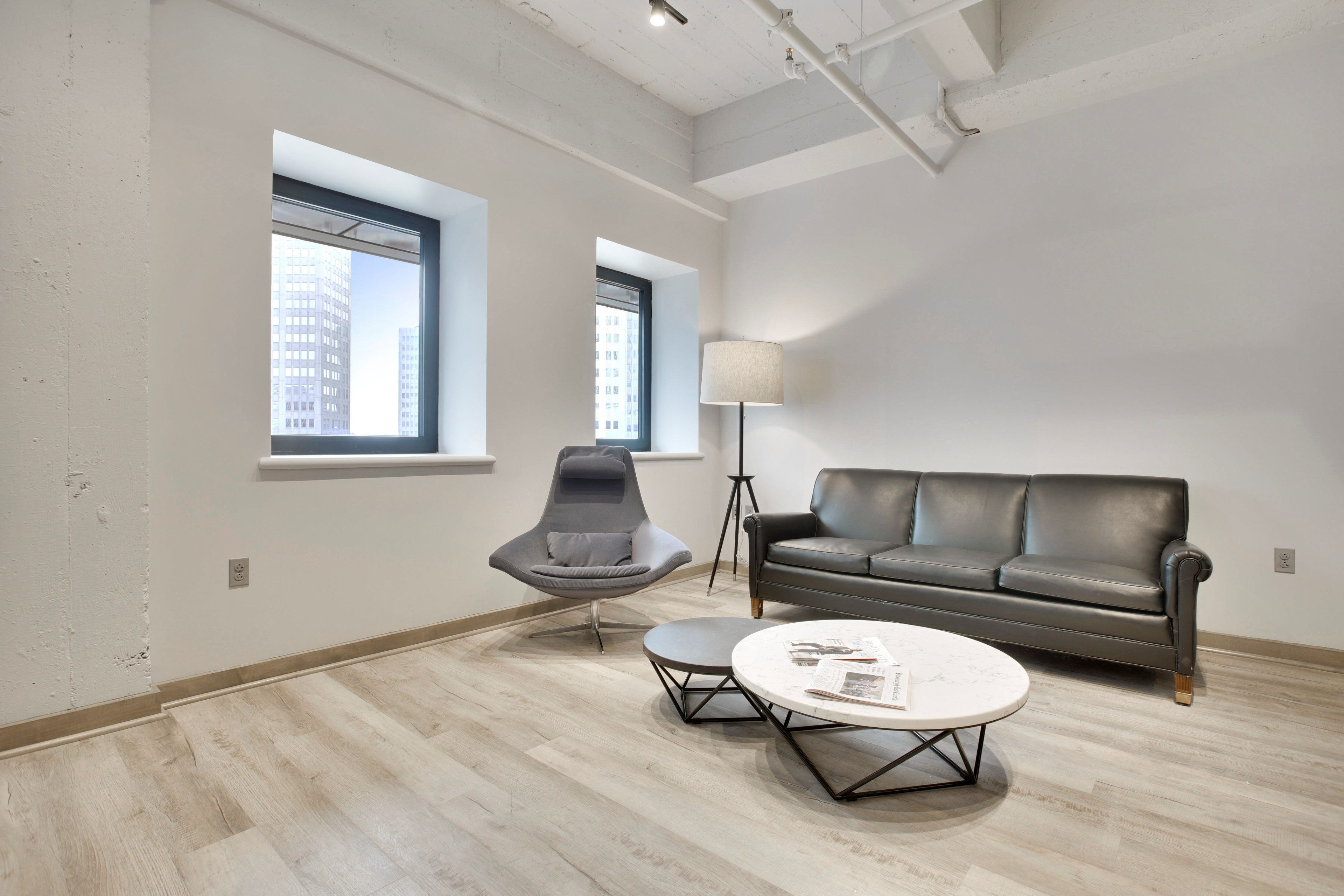 Un salon moderne avec un canapé gris, un fauteuil gris et une table basse ronde. Il y a deux fenêtres qui laissent entrer la lumière naturelle et une lampe sur pied.