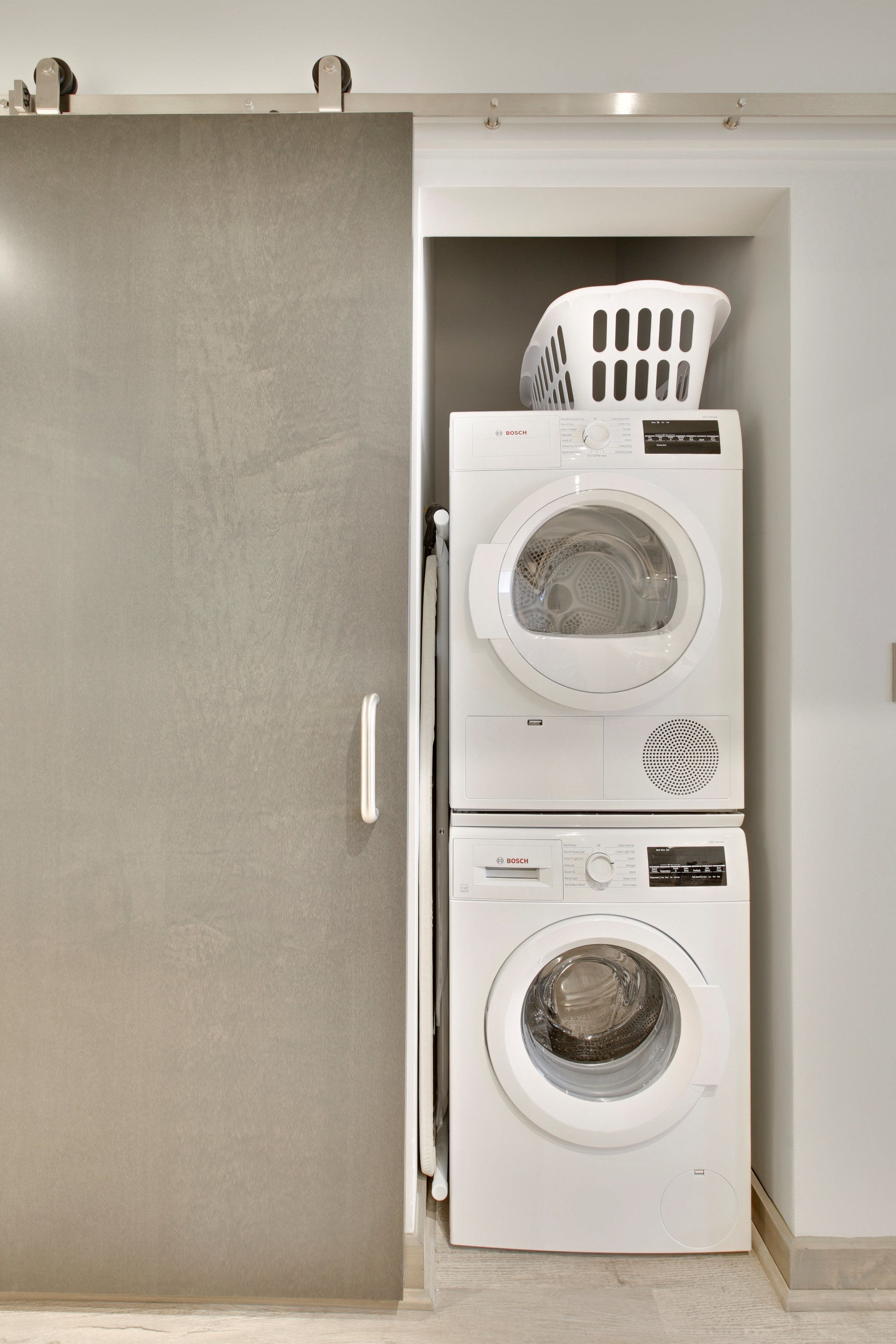A laundry room with two stacked Bosch washing machines and a white plastic laundry basket on top.