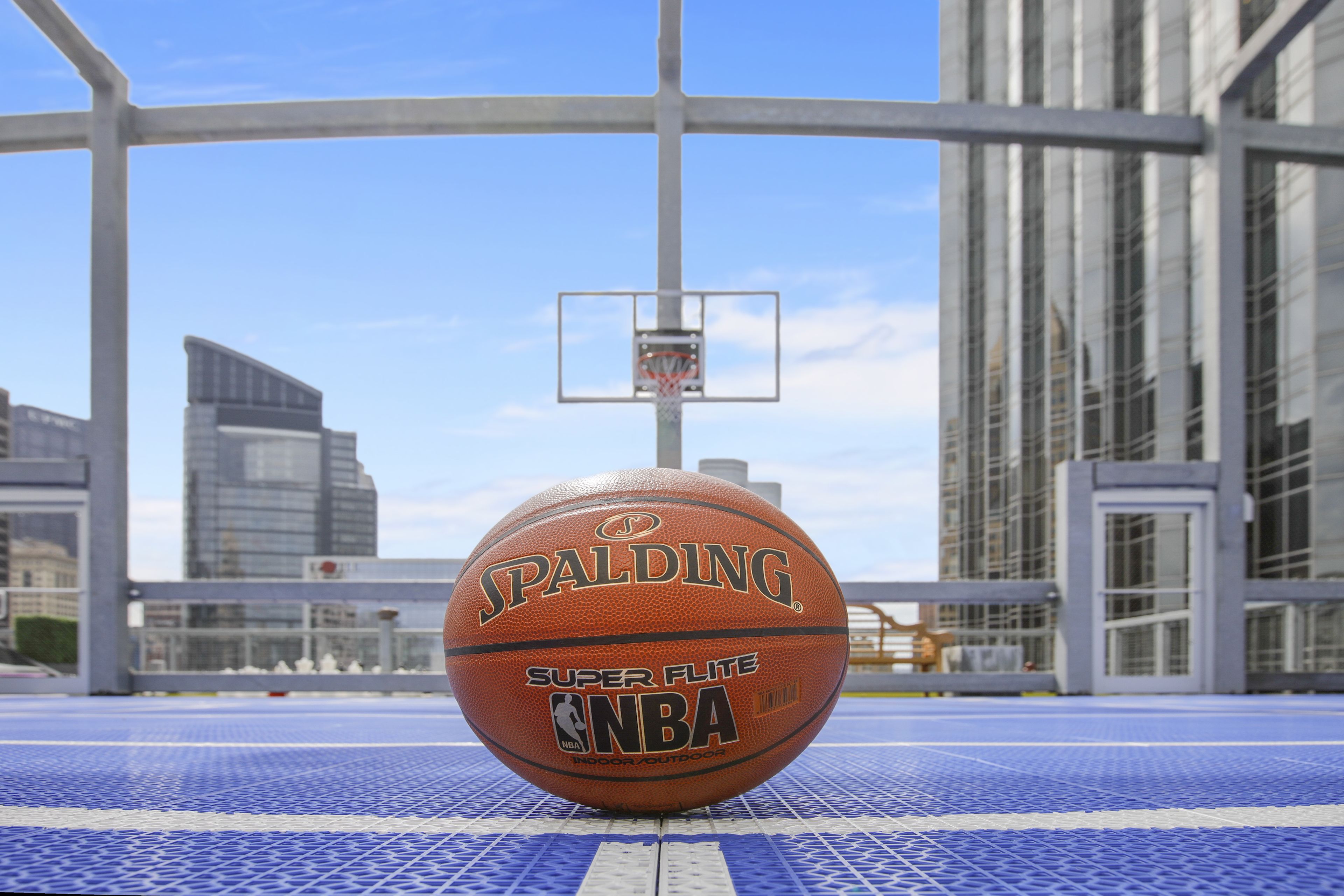Un balón de baloncesto Spalding Super Flite de la NBA sobre una cancha azul al aire libre, con un aro de baloncesto al fondo.