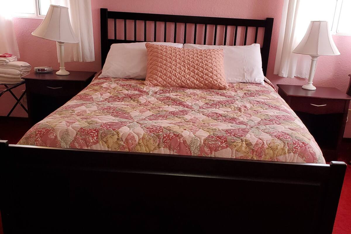 A bed with a floral bedspread, two pillows, a nightstand with a lamp, and a framed picture on the wall.