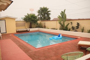 A swimming pool with a blue inflatable float, surrounded by a brick patio, palm trees, and a small building with a door. - Playa Hermosa Inn at the beach (Ensenada)