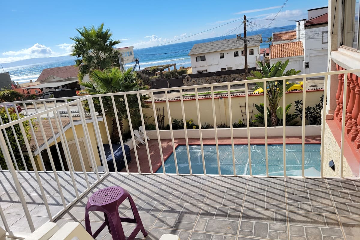 A pool area with a view of the ocean, a white railing, and a purple stool.