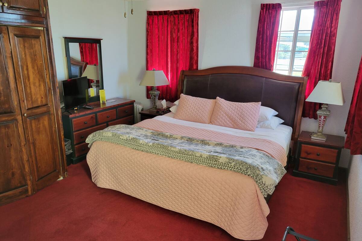 A neatly arranged bedroom with a large bed, wooden headboard, red curtains, and a wooden wardrobe.