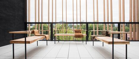 Un espacio exterior moderno con tres bancos de madera y una mesa central, frente a un fondo de listones verticales de madera y vistas de la ciudad.