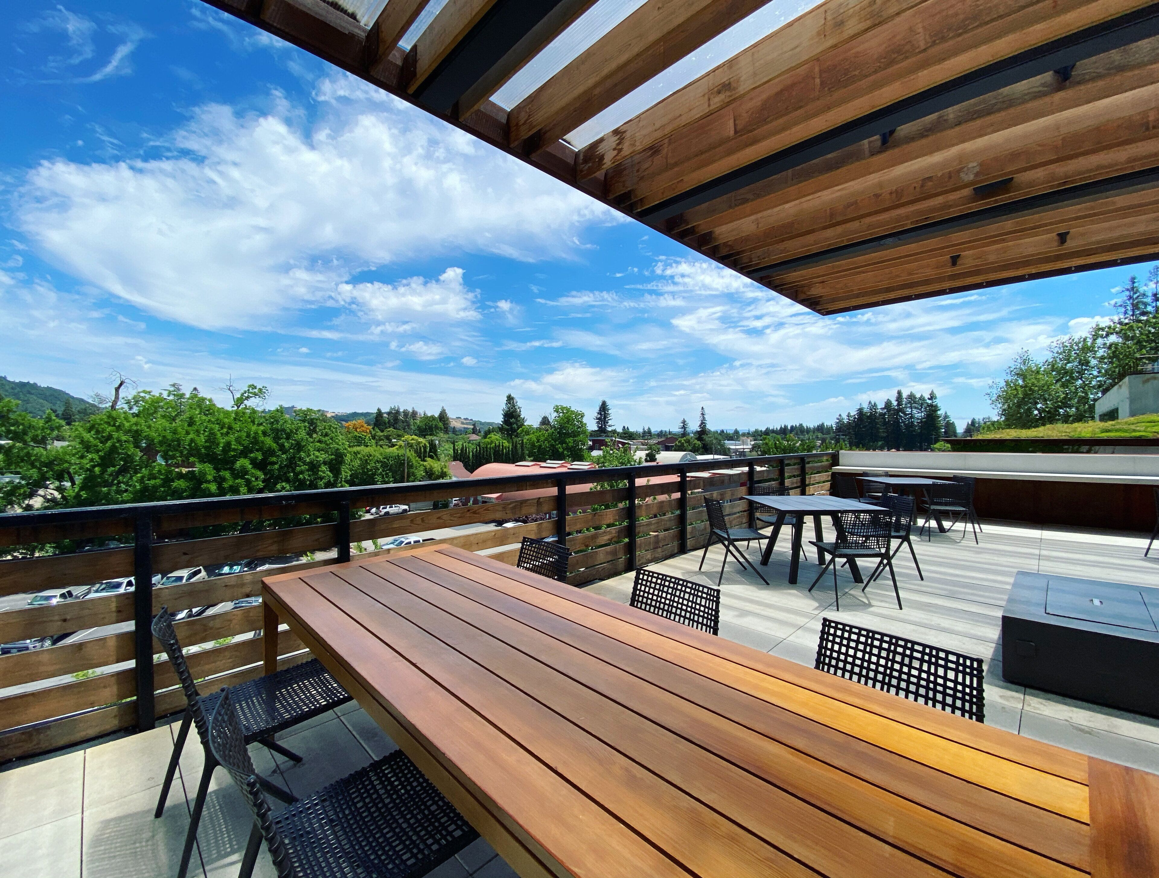 Une terrasse extérieure avec des tables et des chaises en bois, une vue sur un paysage verdoyant et un ciel bleu dégagé.