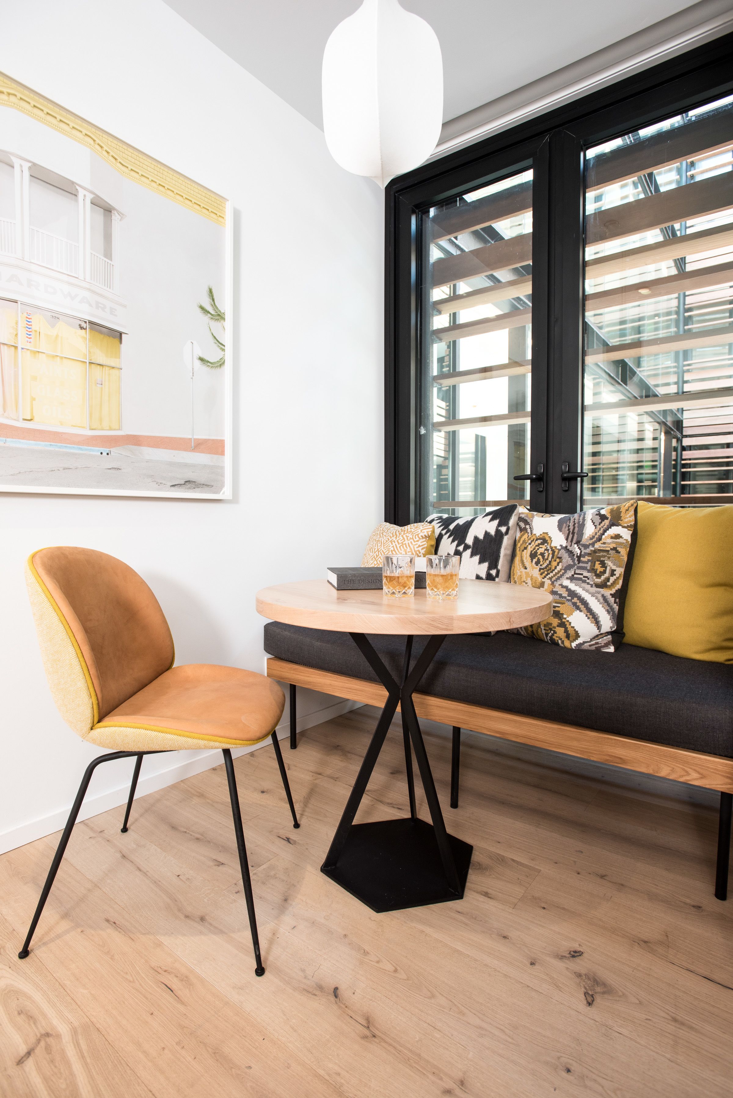 Una sala de estar moderna con una mesa redonda de madera, un sofá con cojines estampados, una sola silla y un ventanal con vistas a un edificio.