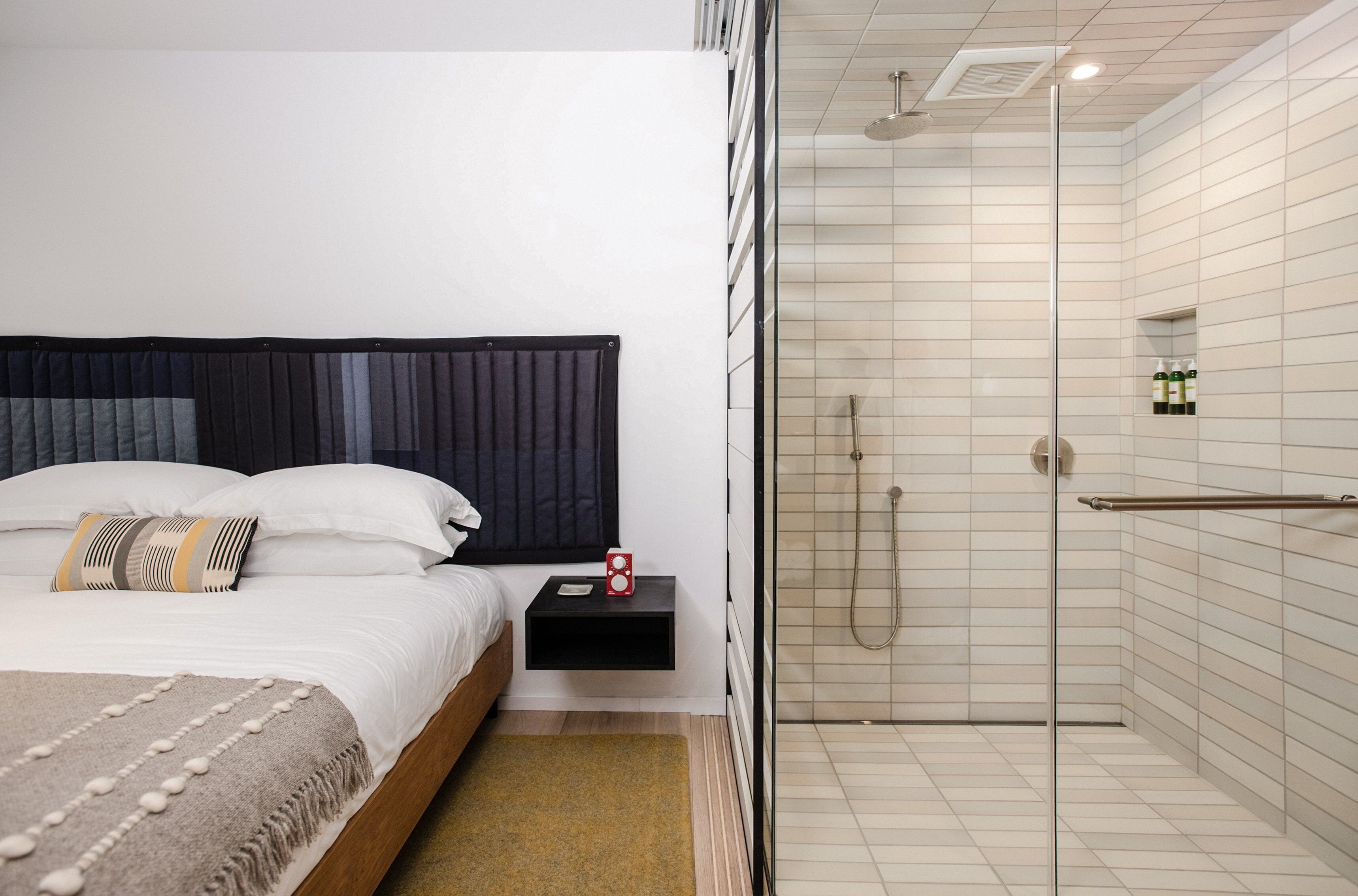 Ein modernes Hotelzimmer mit einem großen Bett, einem Nachttisch mit roter Uhr und einem Glaskabinentrennendes Duschbad.